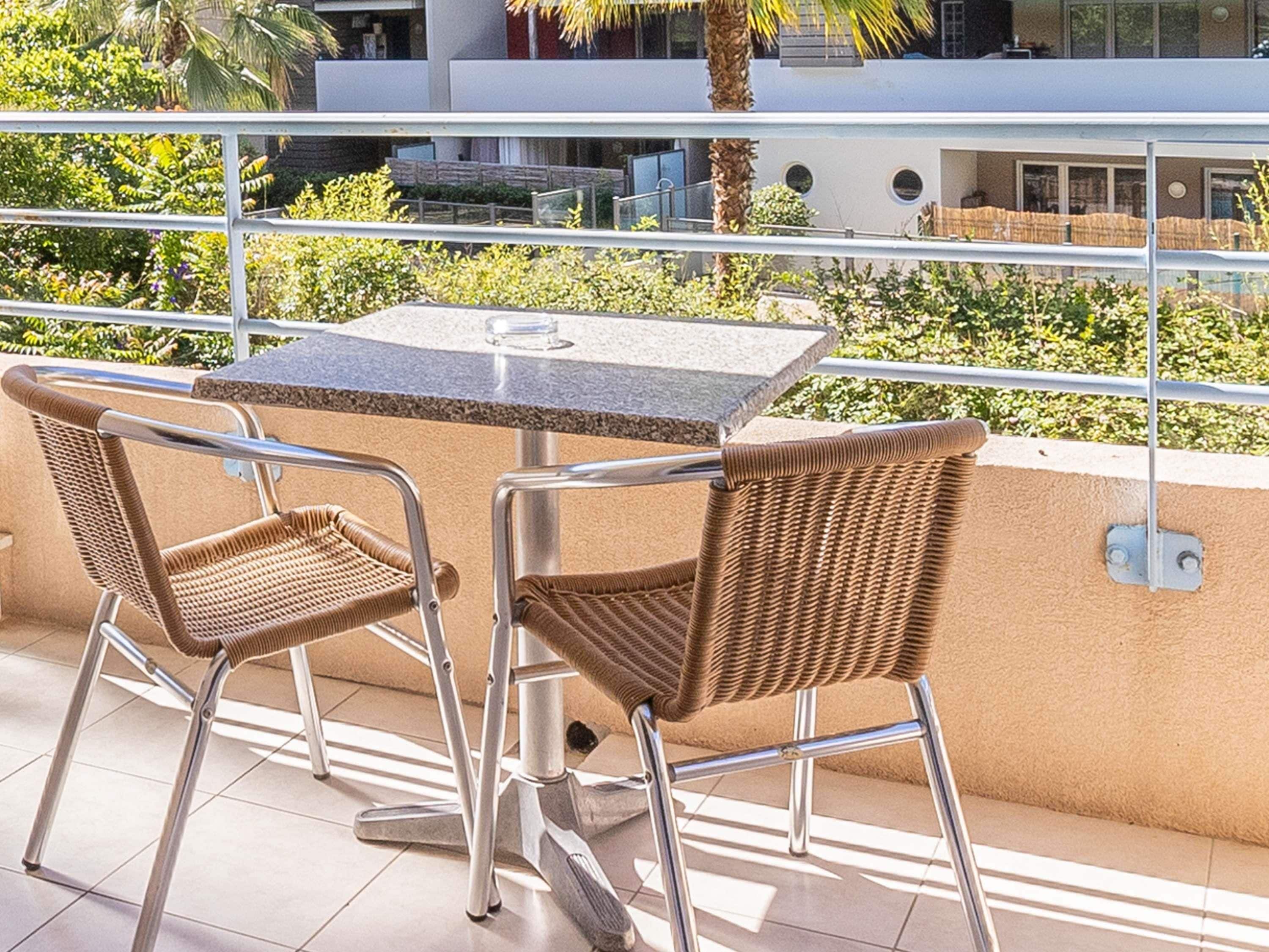 A balcony with a table and two wicker chairs, overlooking a building and greenery.