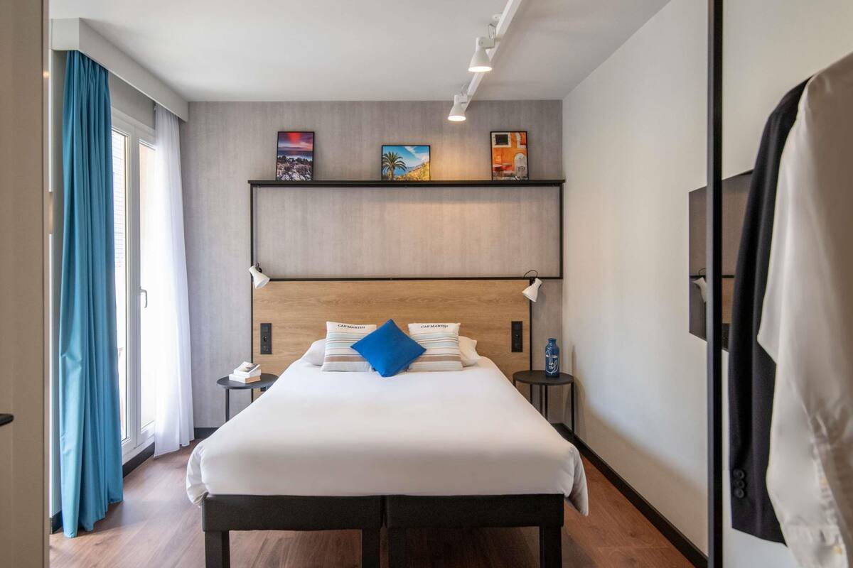 Une chambre d’hôtel avec un grand lit, une table de chevet, une étagère murale décorée d’œuvres d’art et une fenêtre avec des rideaux.
