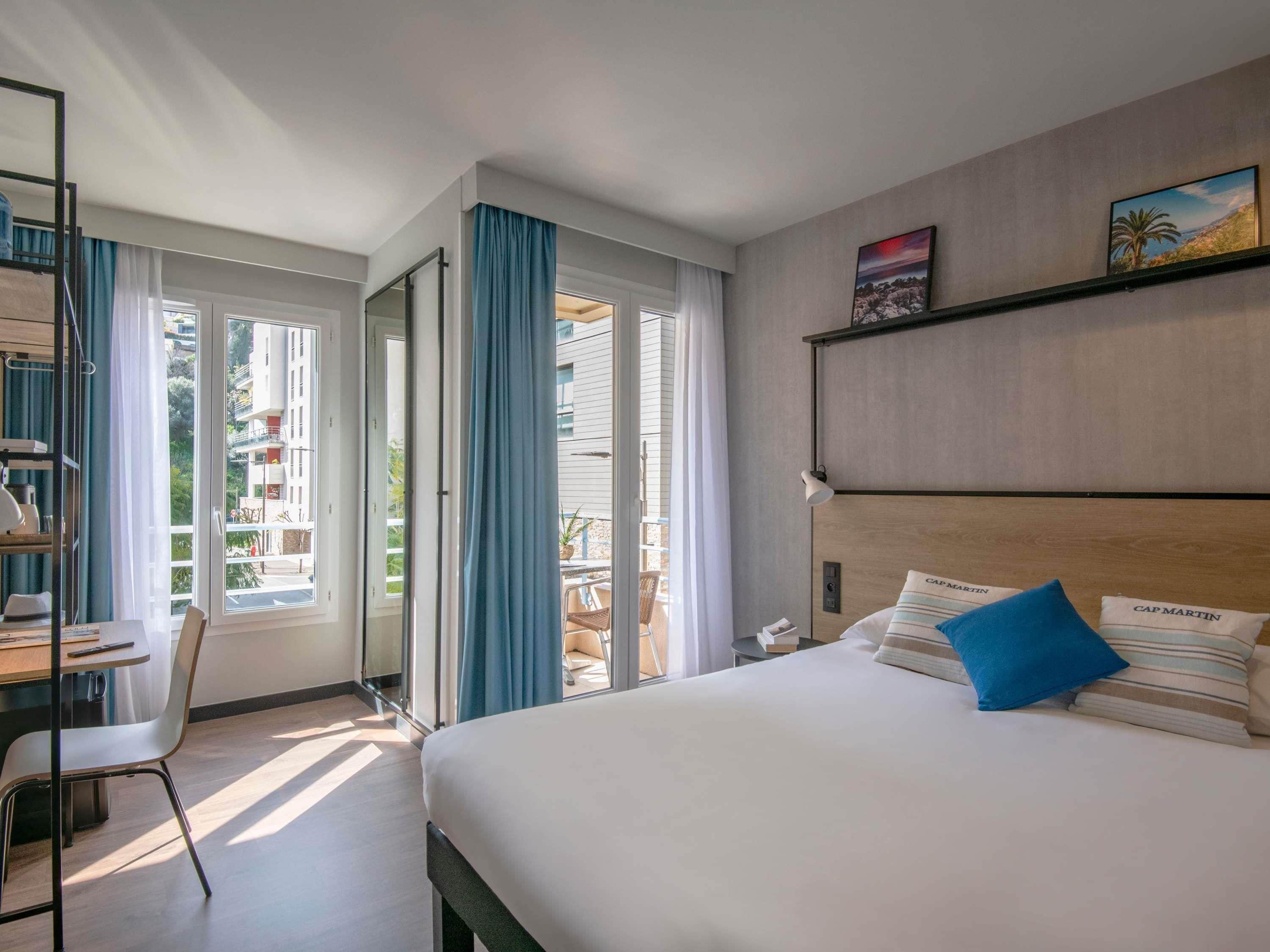 Et hotelværelse med seng, skrivebord og stol. Et vindue med gardiner, et billede på væggen og udsigt ud
