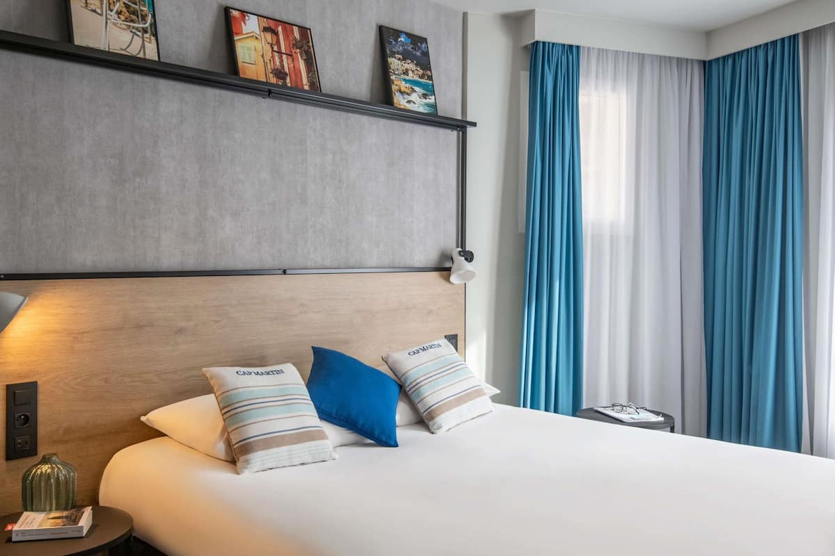 Une chambre d’hôtel avec un grand lit, une tête de lit ornée d’œuvres d’art, une table de chevet avec une lampe et une vue sur la mer à travers les rideaux.