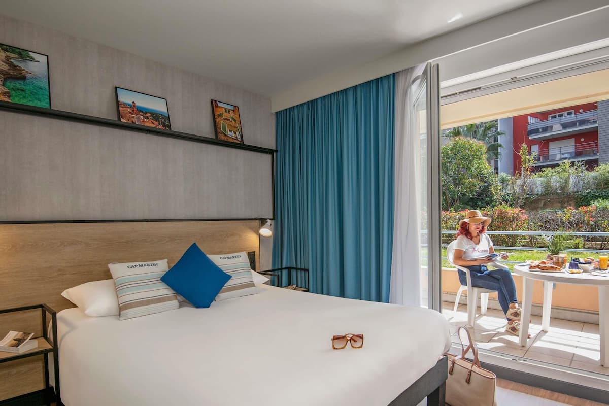 Une chambre d’hôtel avec un grand lit, une table de chevet avec une lampe, une fenêtre avec des rideaux bleus et une vue sur l’extérieur.