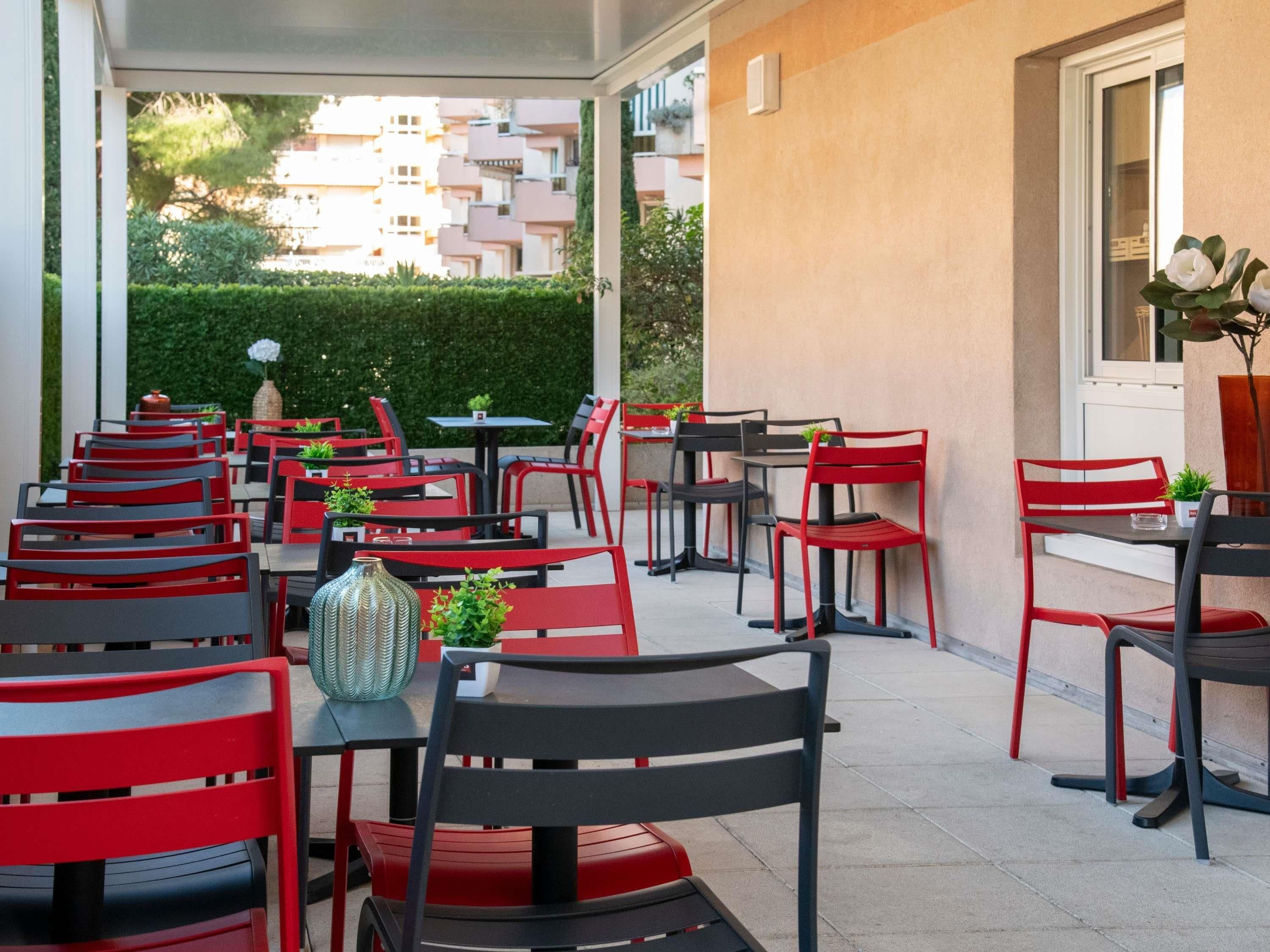 An outdoor cafe area with red and black chairs and tables, a potted plant, and a view of buildings in the background.
