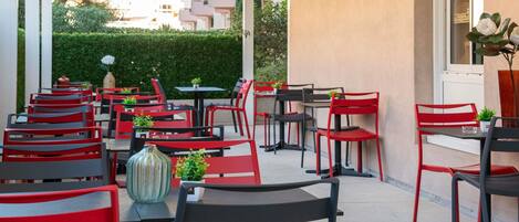An outdoor cafe area with red and black chairs and tables, a potted plant, and a view of buildings in the background.