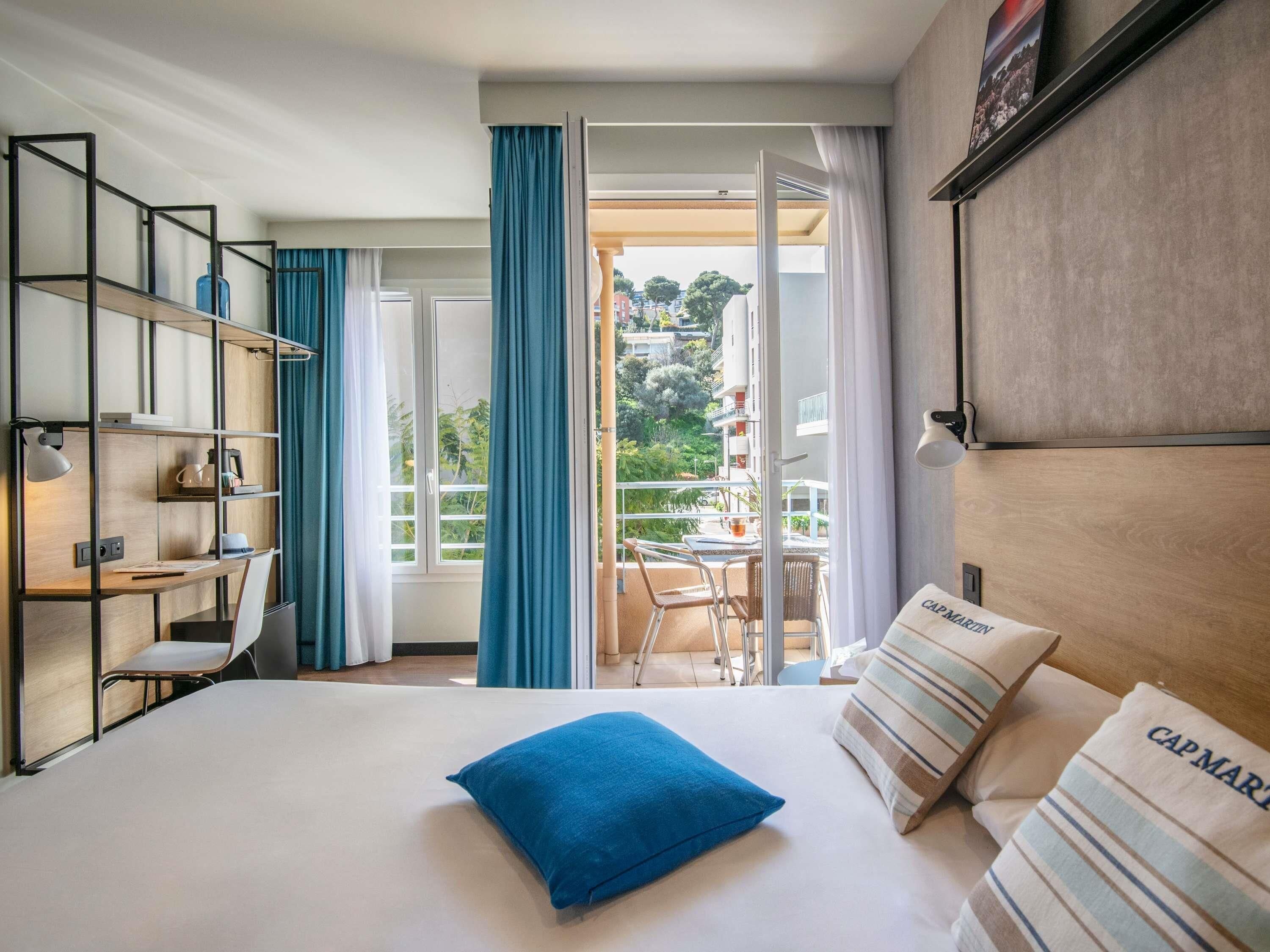 A hotel room with a bed, a desk, a chair, a balcony with a table and chairs, and a view of the outside.