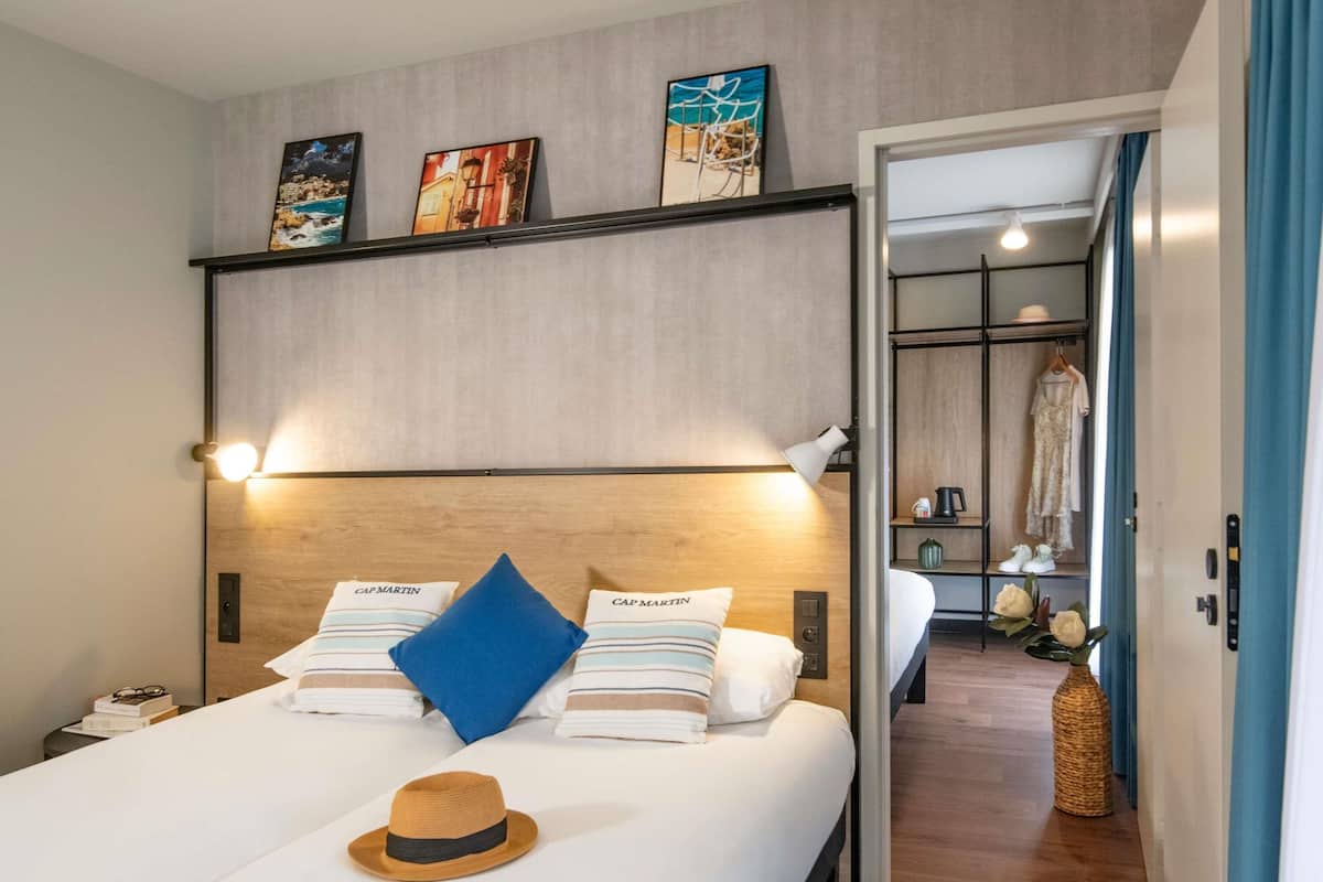 Une chambre d’hôtel avec un lit, une table de chevet, un chapeau et une étagère avec des photos.