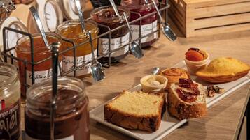 Une variété d’aliments, notamment des pots de confiture, du pain et des pâtisseries, sont disposés sur une table en bois.