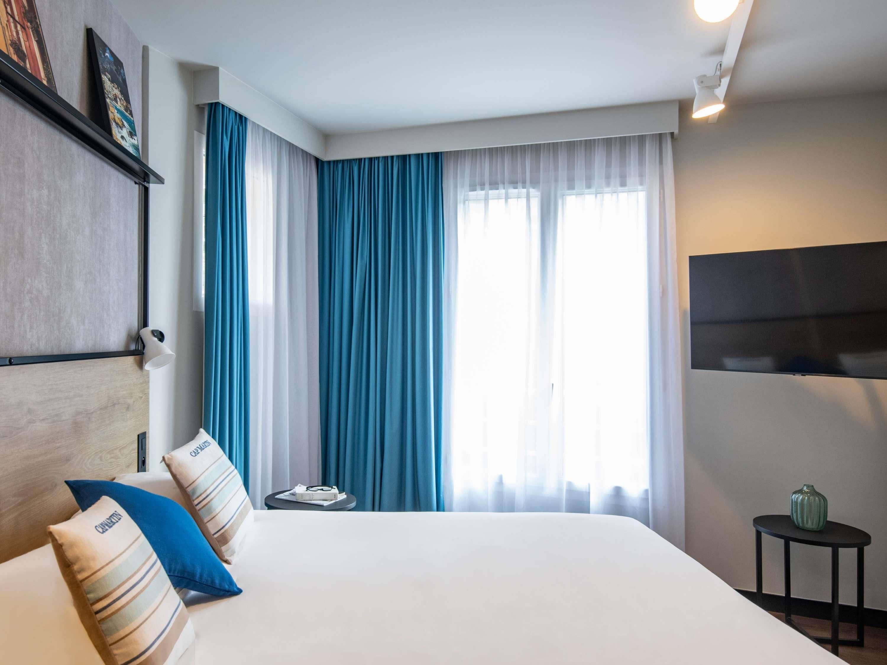 A hotel room with a large bed, a flat-screen TV, a small table with a vase, and a window with blue curtains.