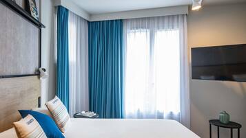 A hotel room with a large bed, a flat-screen TV, a small table with a vase, and a window with blue curtains.