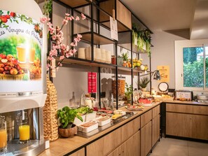 Un bufé de desayuno con diversos alimentos, un dispensador de zumo y plantas decorativas.
