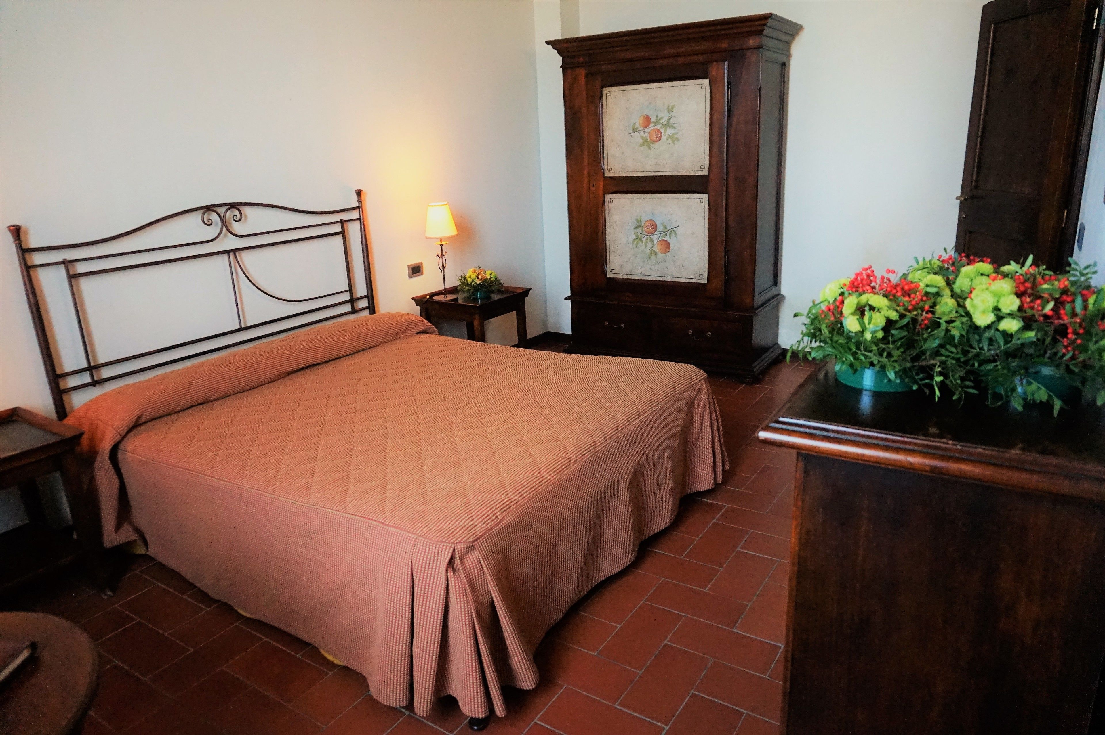 A neatly made bed with a headboard, a bedside table with a lamp, a wooden cabinet with two framed pictures, and a potted plant with red berries.