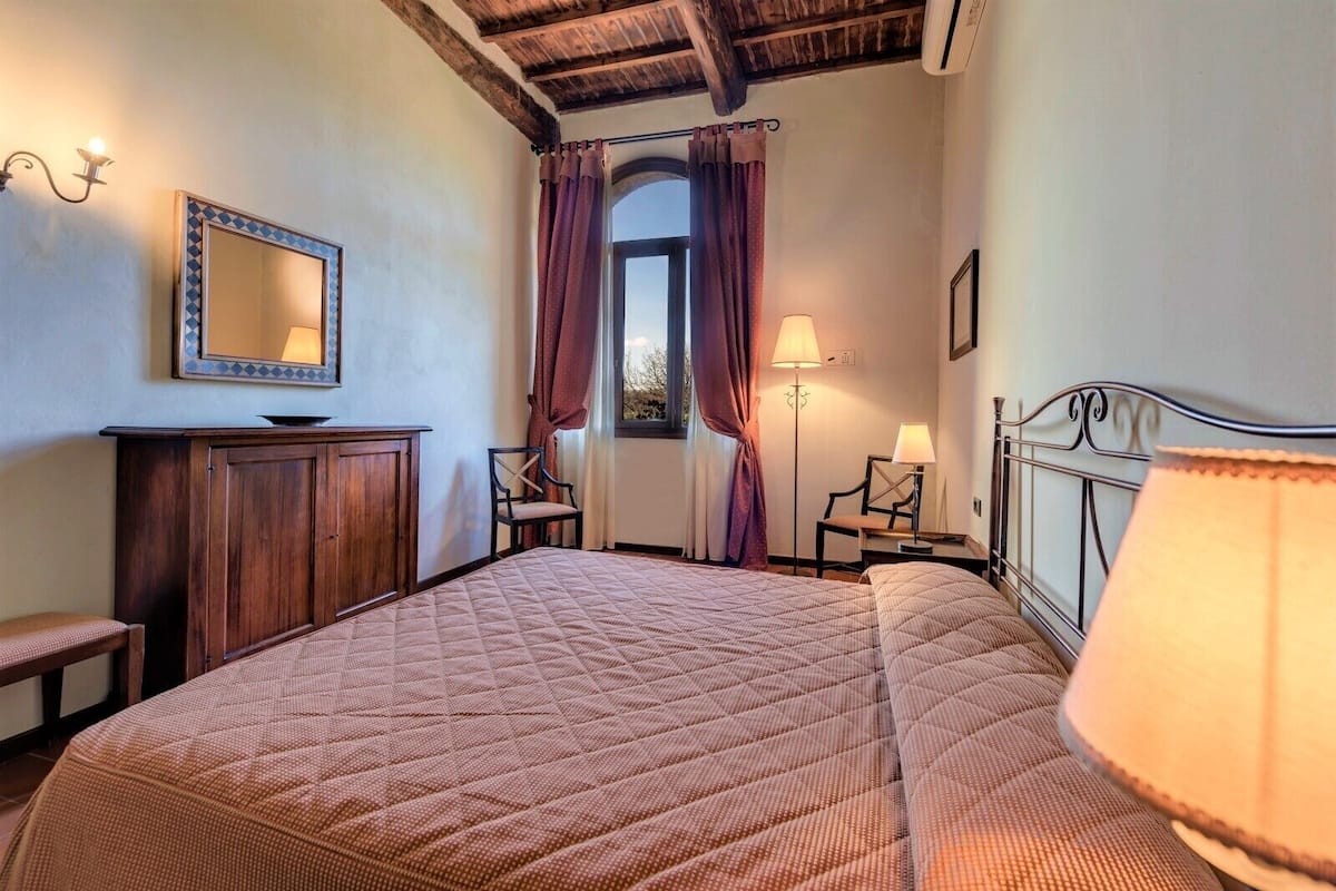 A bedroom with a large bed, a wooden dresser, a chair, a desk with a lamp, and a window with curtains.