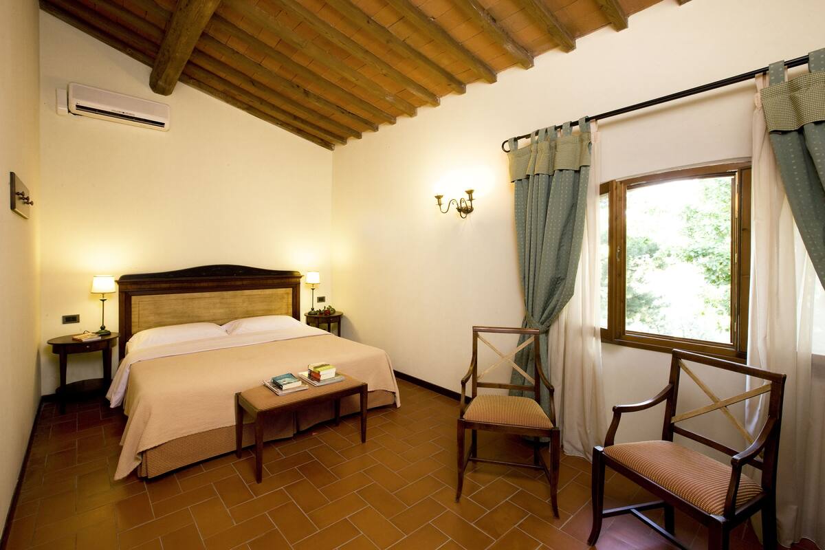 A hotel room with a wooden ceiling, a bed with a headboard, a bedside table with a lamp, a chair, and a window with curtains.