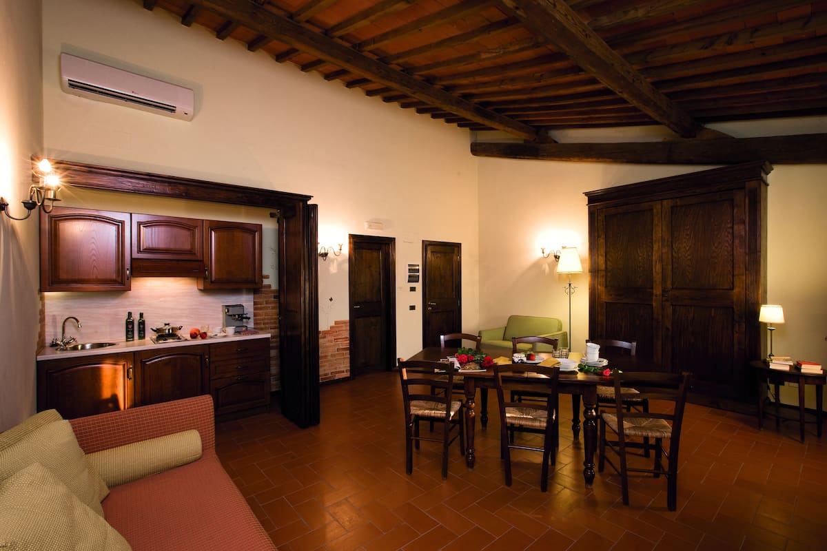 A cozy dining area with a wooden table and chairs, a kitchen with wooden cabinets, and a brick wall.
