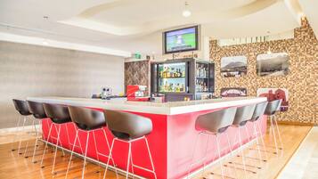 Una zona de bar con mostrador rojo, taburetes negros, un televisor montado sobre la barra y una pared de azulejos con cuadros.