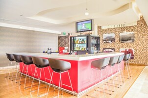 Una zona de bar con mostrador rojo, taburetes negros, un televisor montado sobre la barra y una pared de azulejos con cuadros.