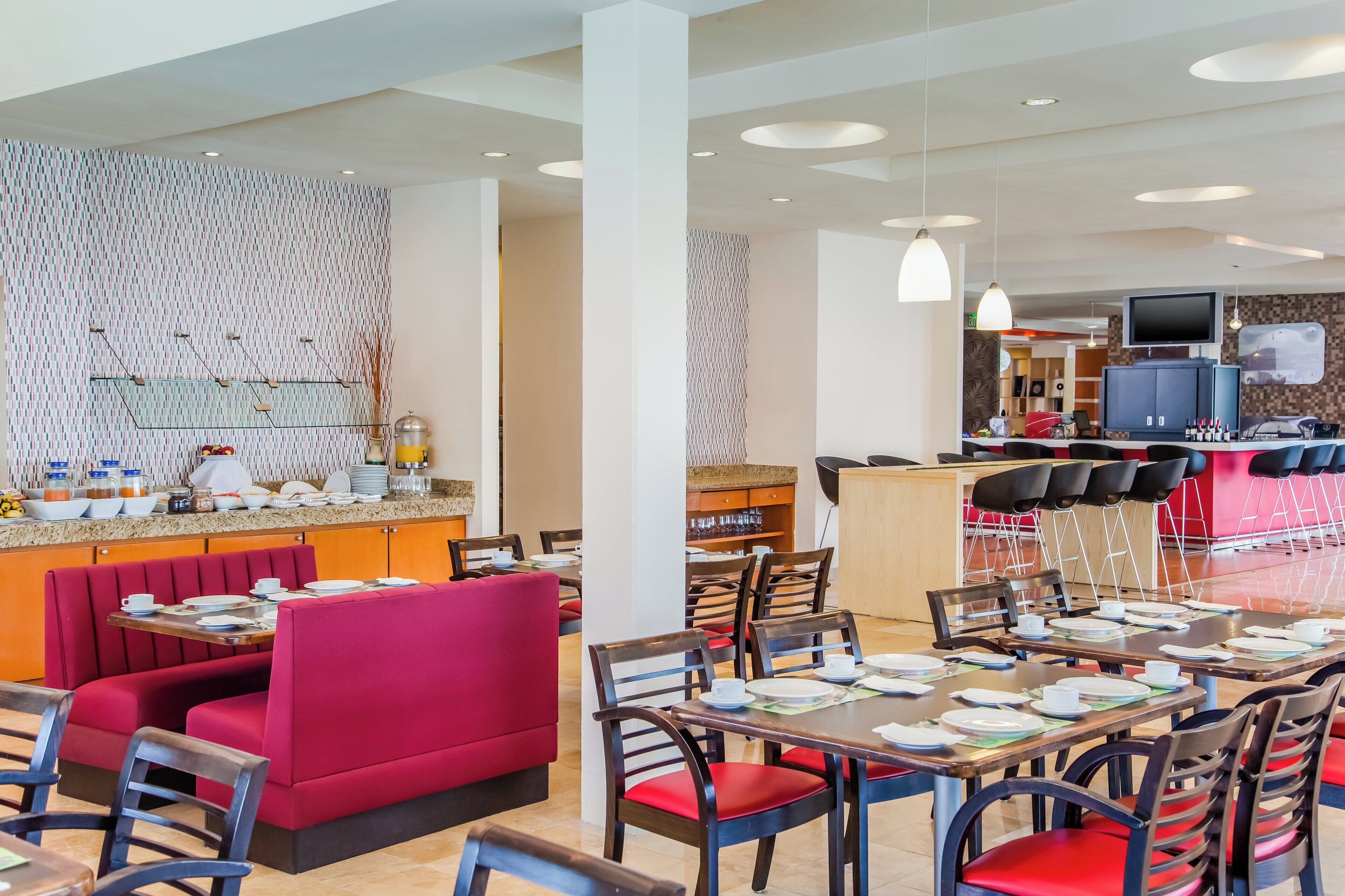 Um espaço de refeições moderno em hotel, com assentos em camarim vermelho e mesas de madeira, uma cozinha ao fundo e uma TV de tela plana na parede.