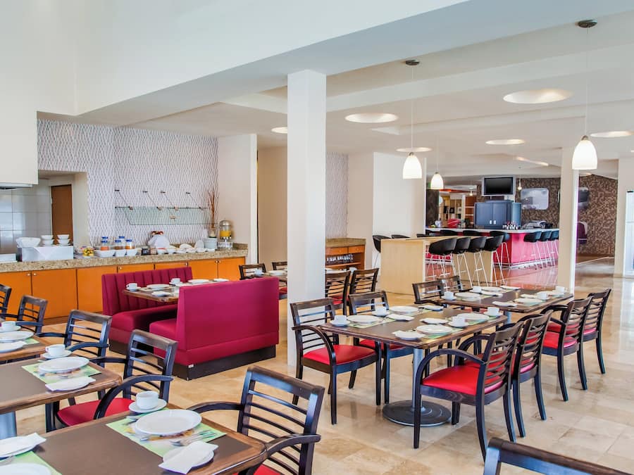 Un comedor de hotel con varias mesas y sillas, una zona de asientos tipo camarote en rojo, y una cocina al fondo.