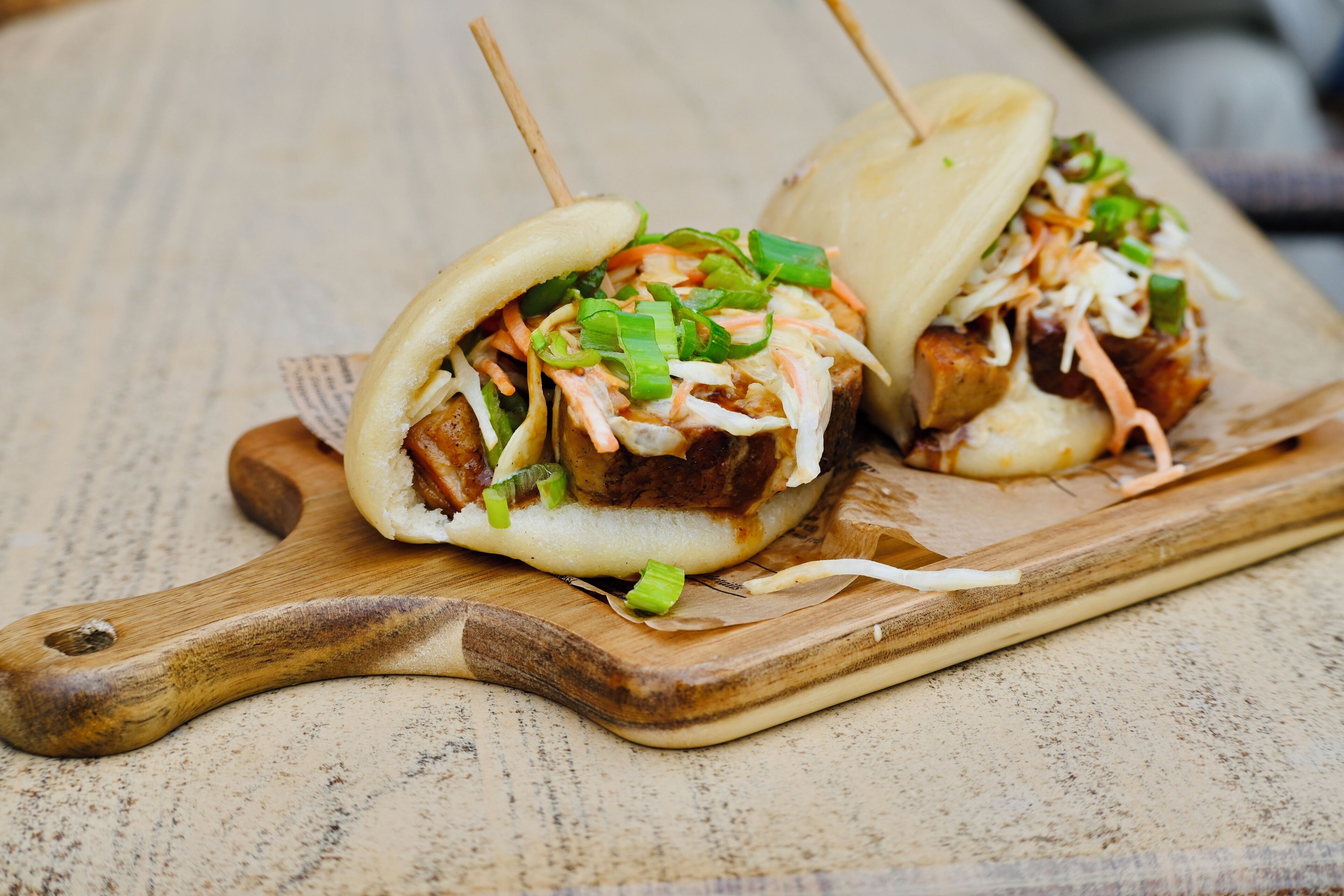 Two bao buns with meat and vegetables on a wooden cutting board.