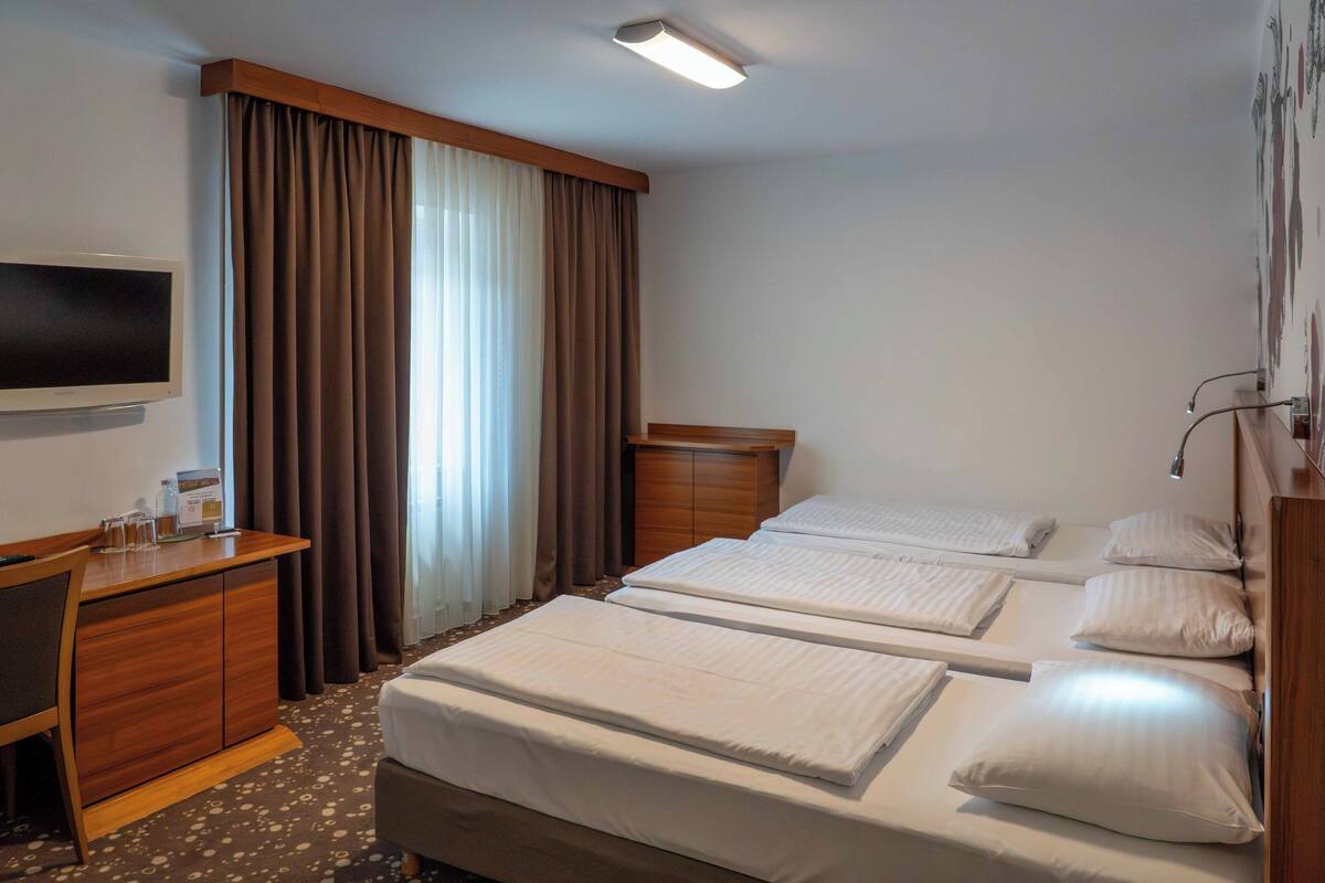 Ein Hotelzimmer mit Schreibtisch, zwei Betten, einem Fernseher und einem großen Fenster mit Vorhängen.