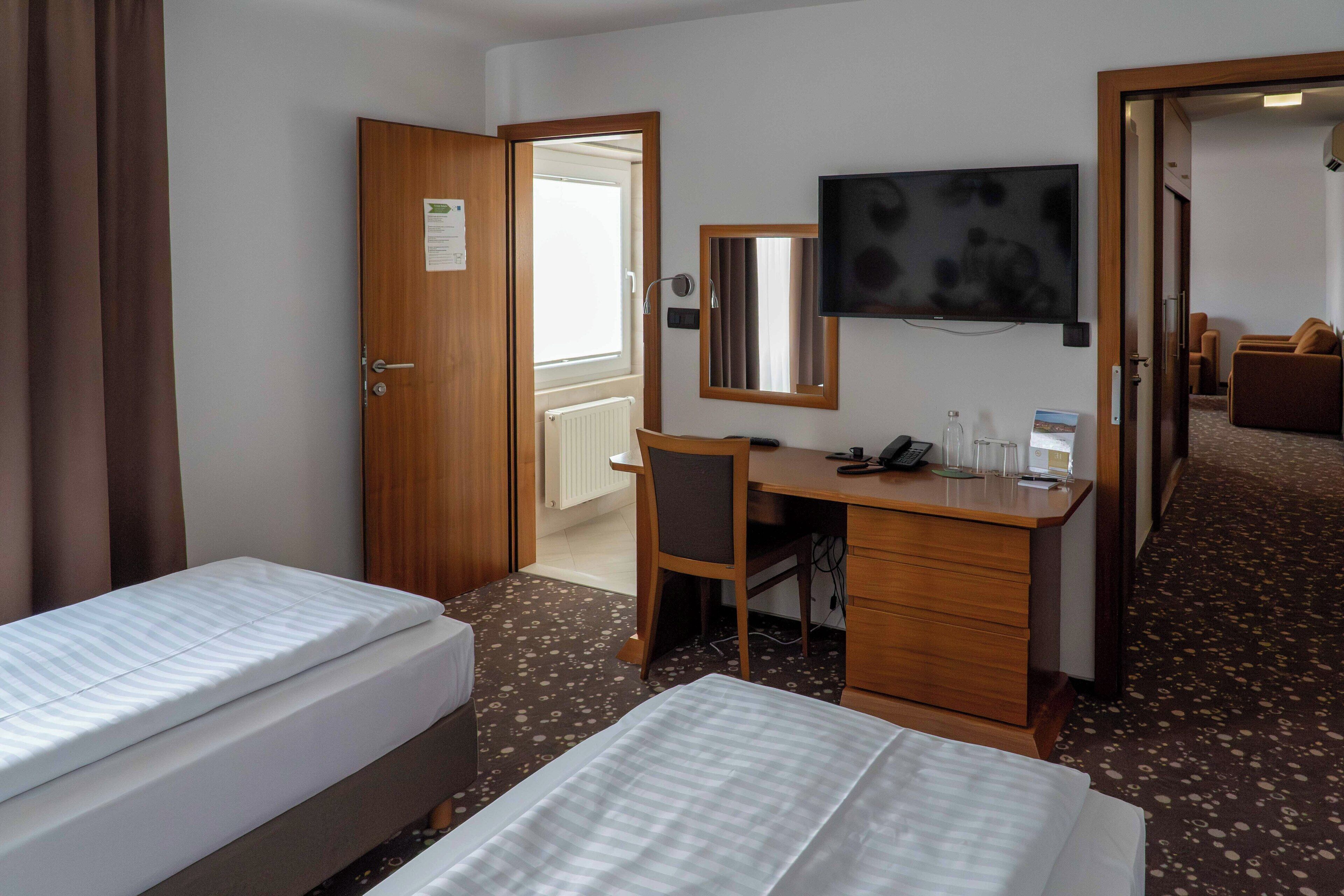 A hotel room with two beds, a desk, a chair, a television, and a bathroom visible through an open door.