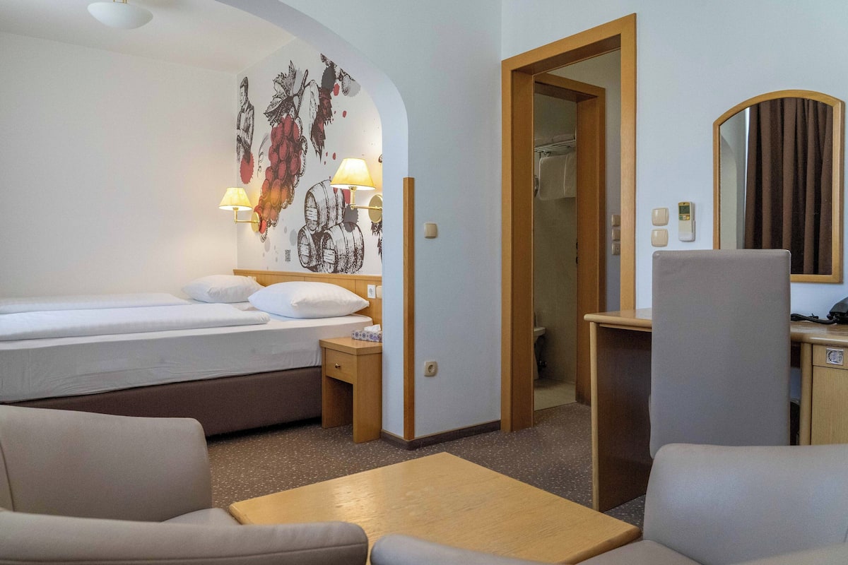 Ein Hotelzimmer mit einem Bett, einem Schreibtisch, einem Stuhl, einem kleinen Tisch, einer Lampe, einem Spiegel und einer Wand mit einem Wandgemälde.