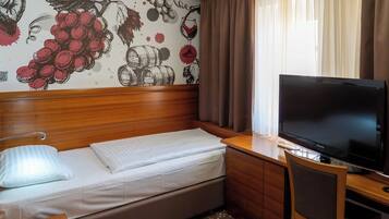 Habitación de hotel compacta con cama, escritorio con televisor e ilustraciones de uvas y vino en la pared.