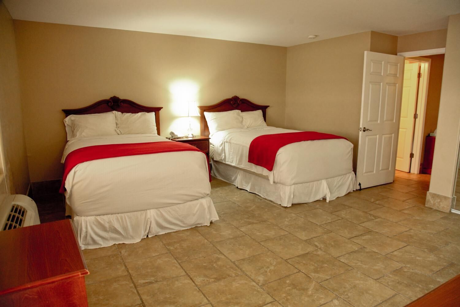 A hotel room with two beds, a nightstand with a lamp, a wooden desk, and a door leading to another room.