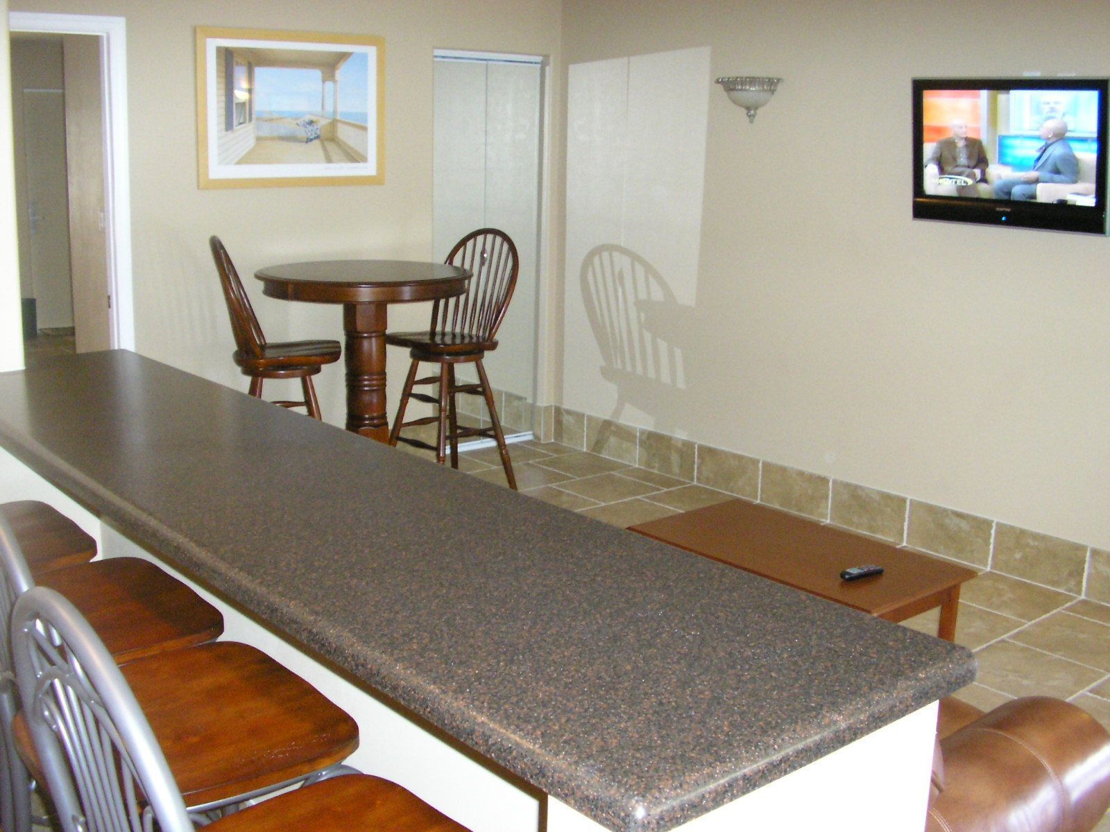 A kitchen with a granite countertop, wooden chairs, a dining table, and a television mounted on the wall.