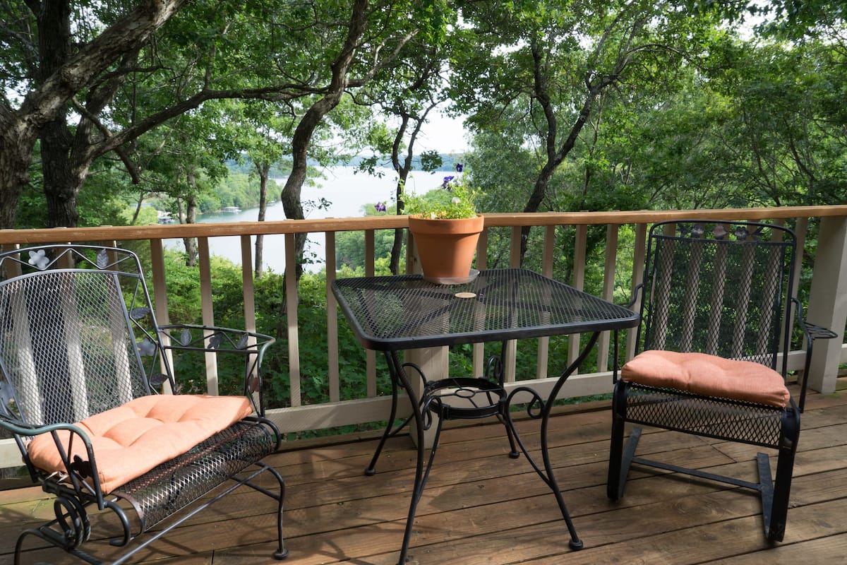 A wooden deck with a metal table and two chairs, overlooking a body of water and surrounded by trees.