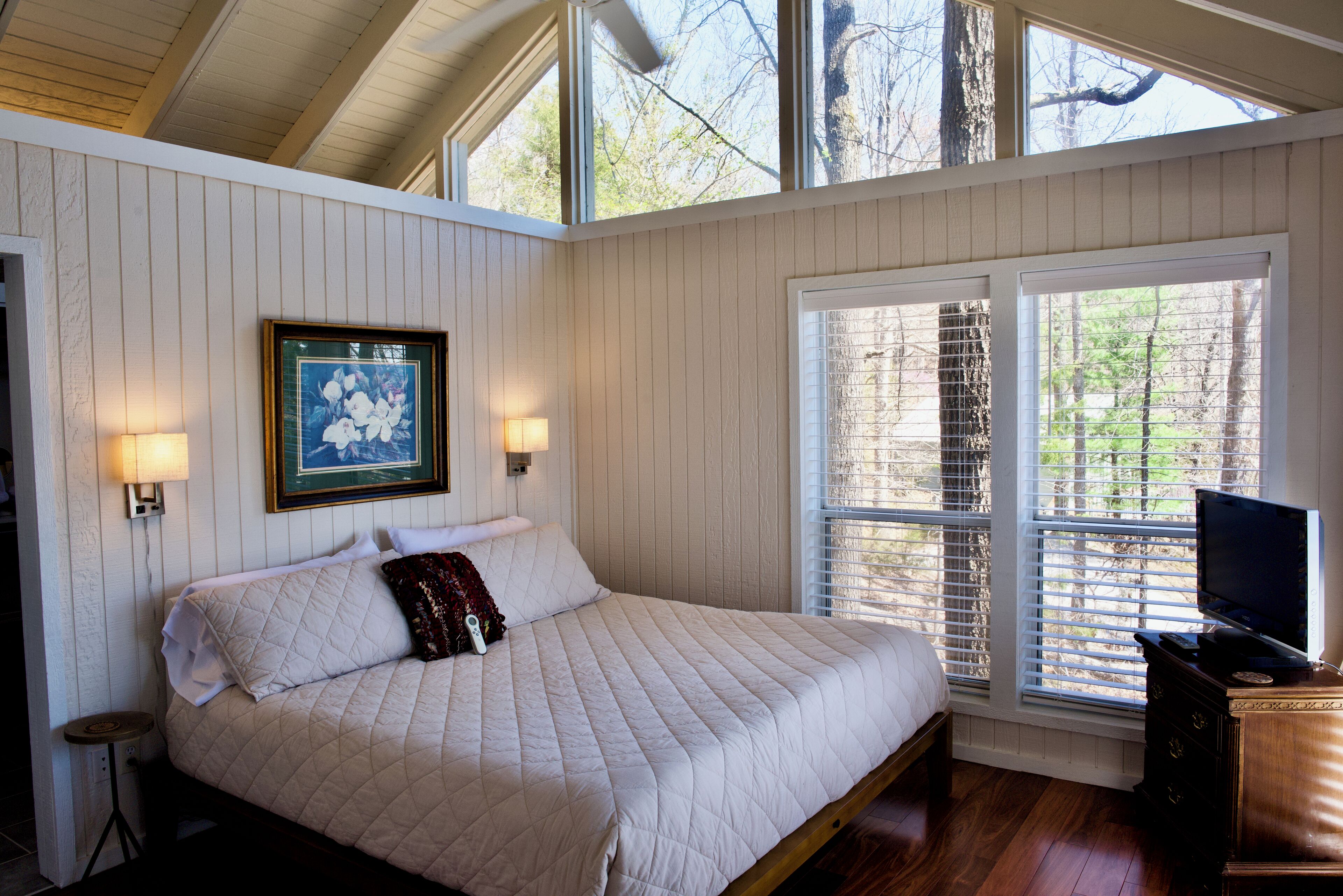 A bedroom with a large bed, two wall-mounted lamps, a framed picture, and a TV on a wooden dresser.