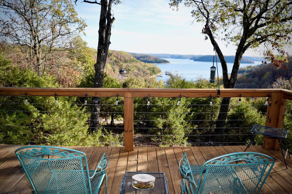 A wooden deck with two turquoise chairs and a table, overlooking a lake and hills.