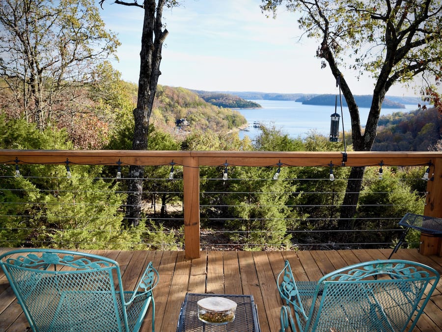 Una terraza de madera con dos sillas turquesas y una mesa, con vista a un lago y colinas.