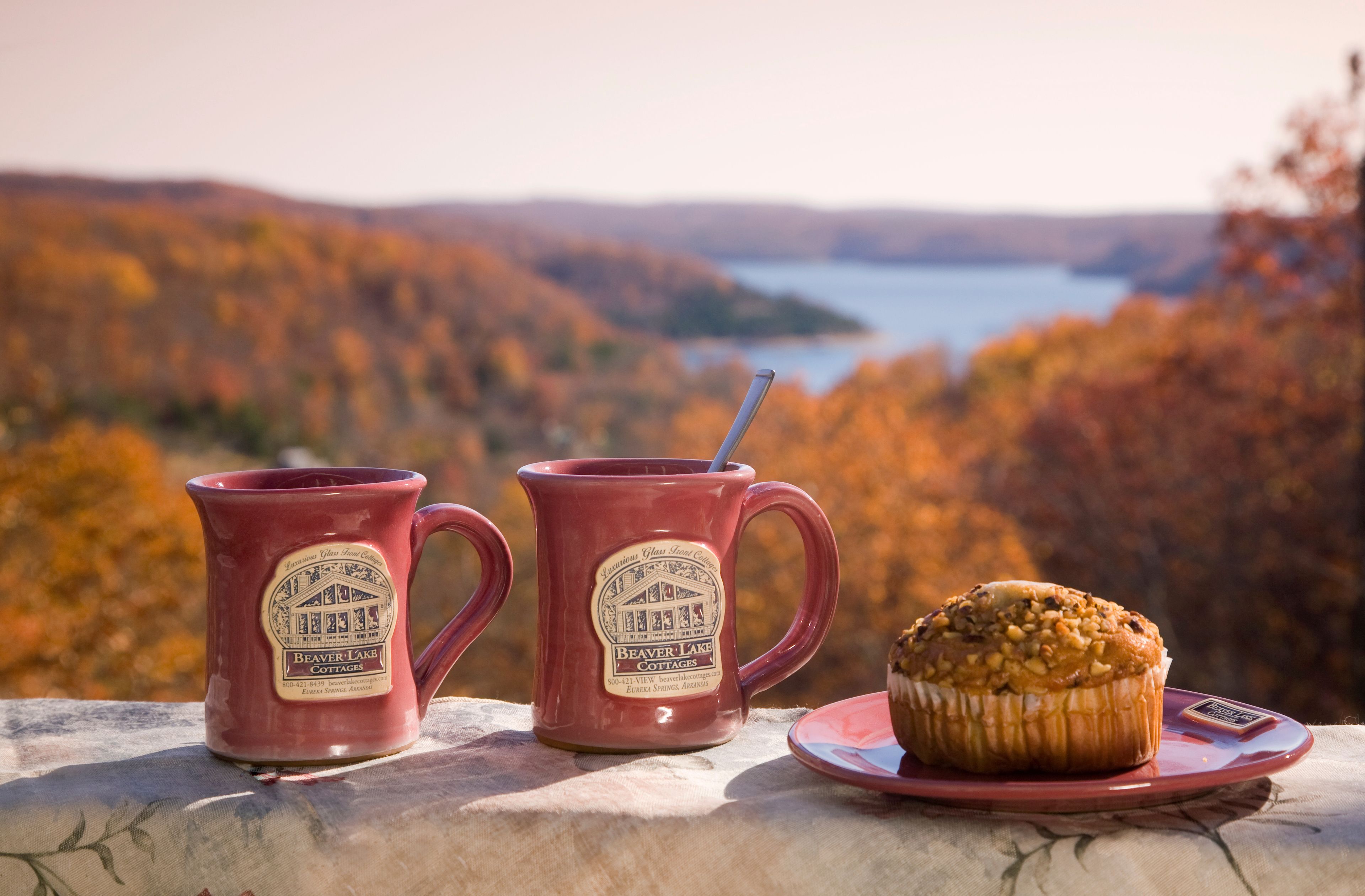 Dos tazas con etiqueta, un muffin en un plato, frente a una vista panorámica de un lago y follaje otoñal.