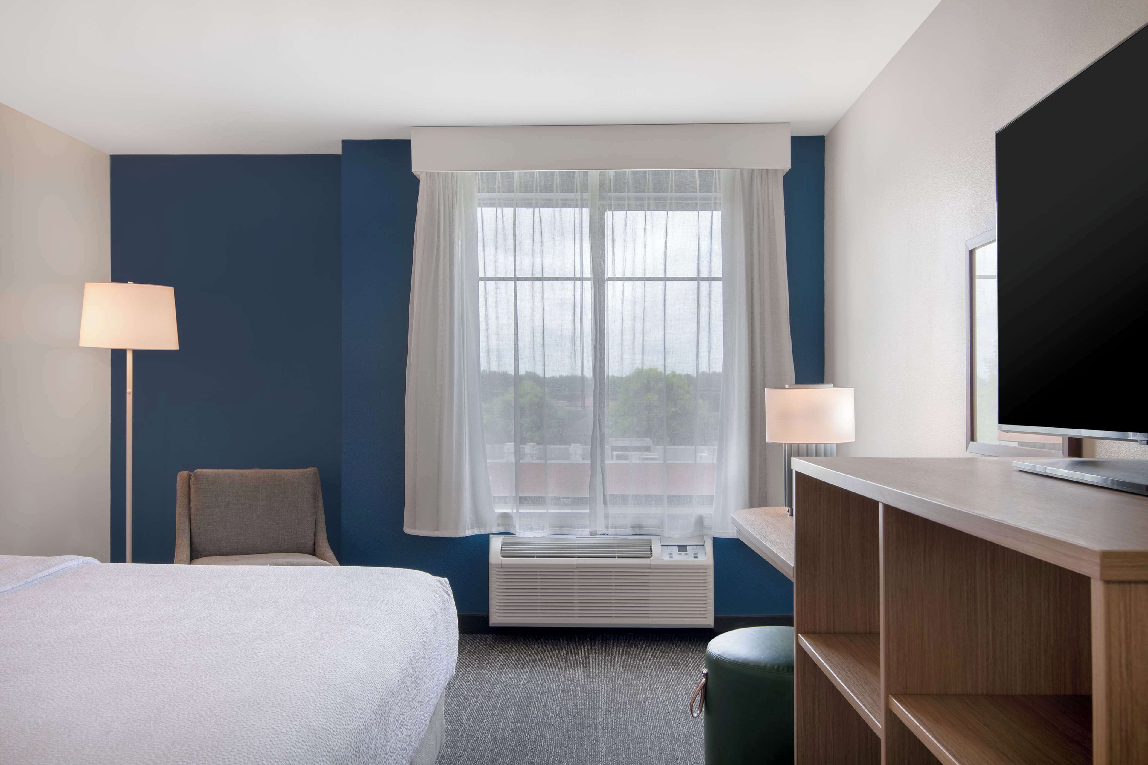 A hotel room with a bed, a chair, a desk with a TV, and a window with curtains.
