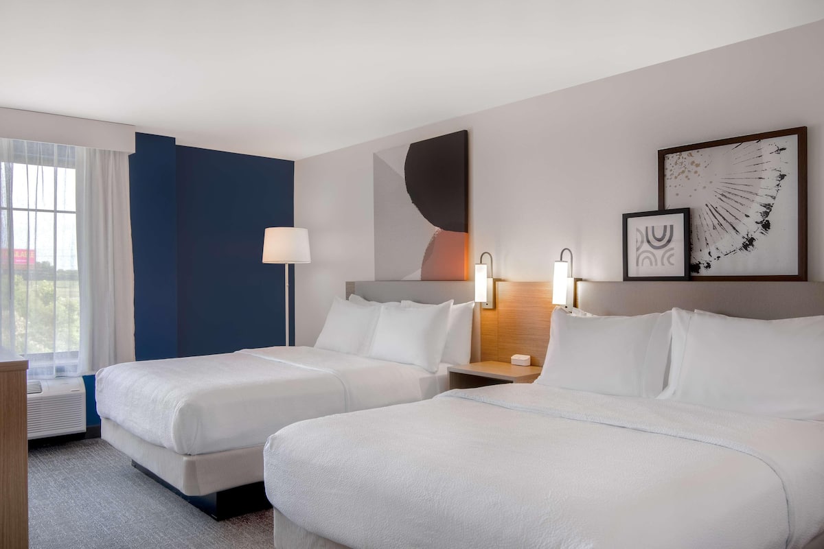 A hotel room with two beds, a nightstand, a lamp, and artwork on the wall.