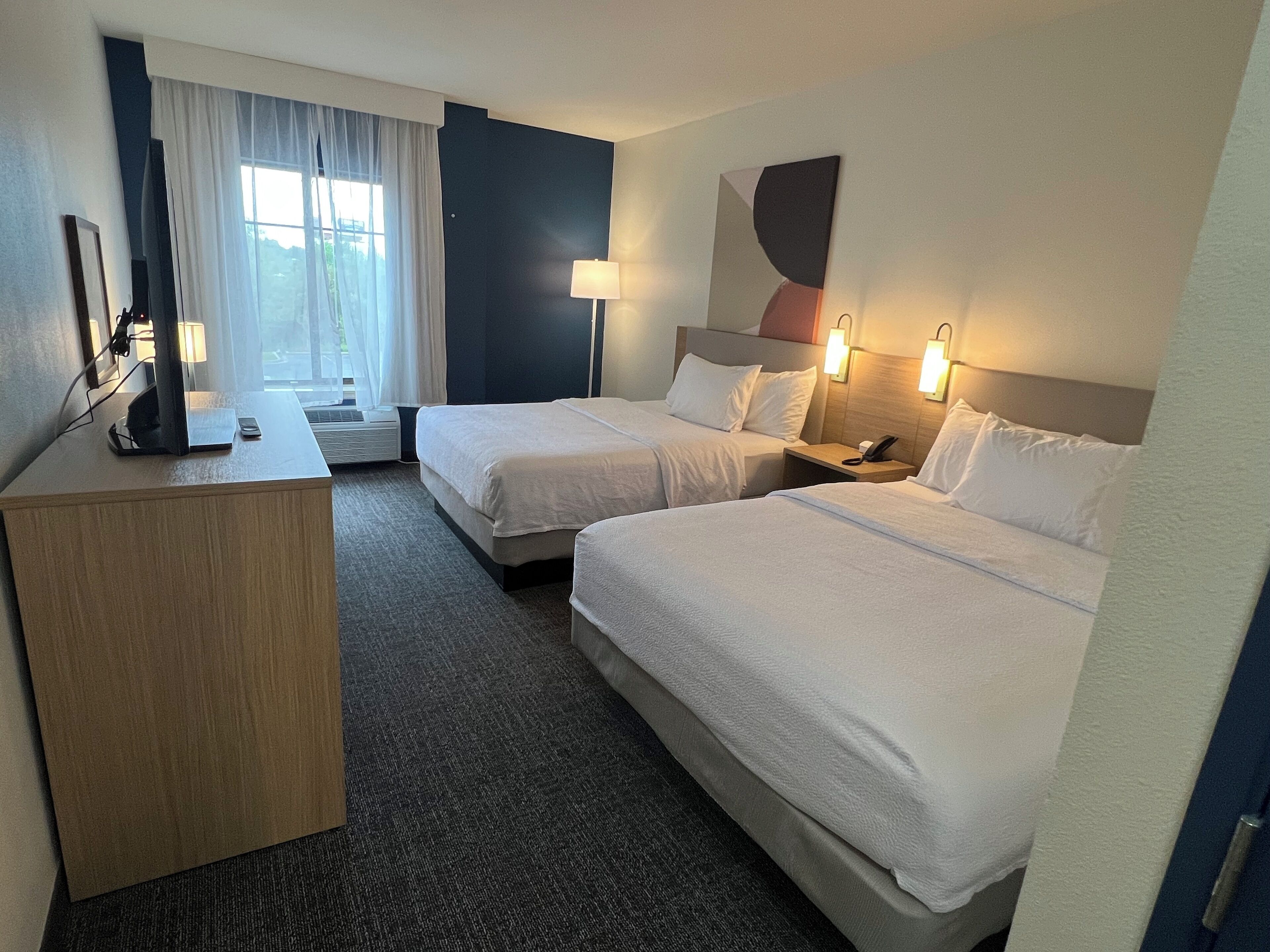 Habitación de hotel con dos camas, un escritorio, una lámpara y un ventanal grande con cortinas.