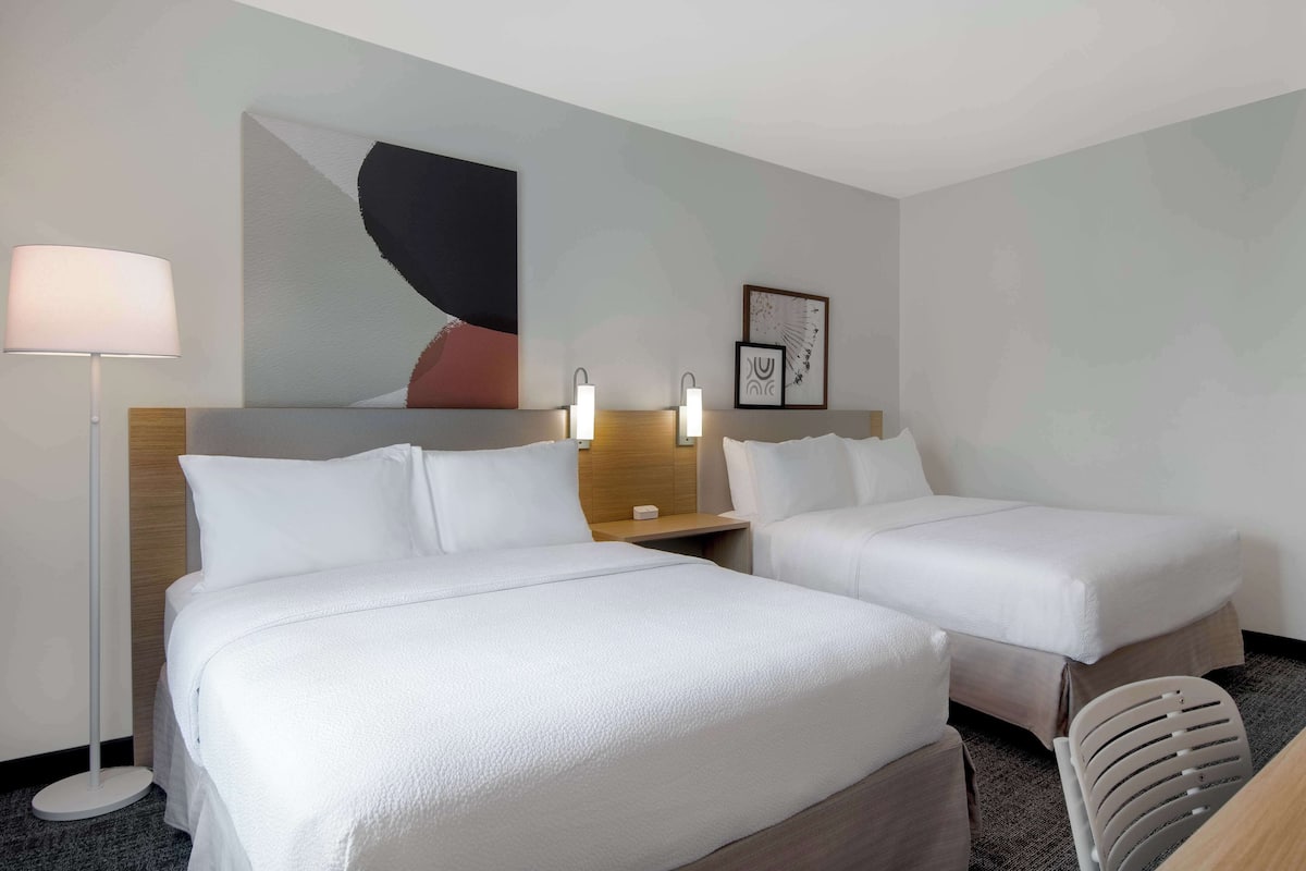 A hotel room with two beds, a desk, a chair, a lamp, and wall art.