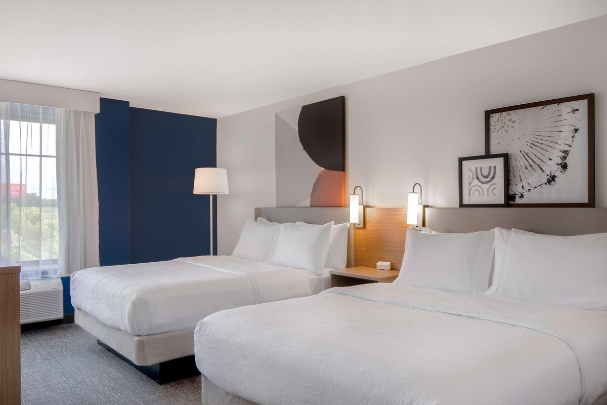 A hotel room with two beds, a nightstand, a lamp, and artwork on the wall.