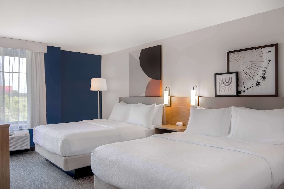 A hotel room with two beds, a nightstand, a lamp, and artwork on the wall.
