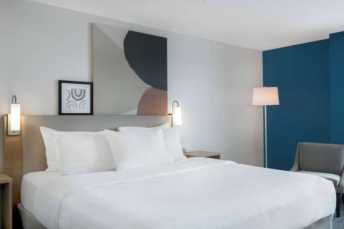 A hotel room with a large bed, two bedside tables, a chair, a wall-mounted lamp, and a framed picture.