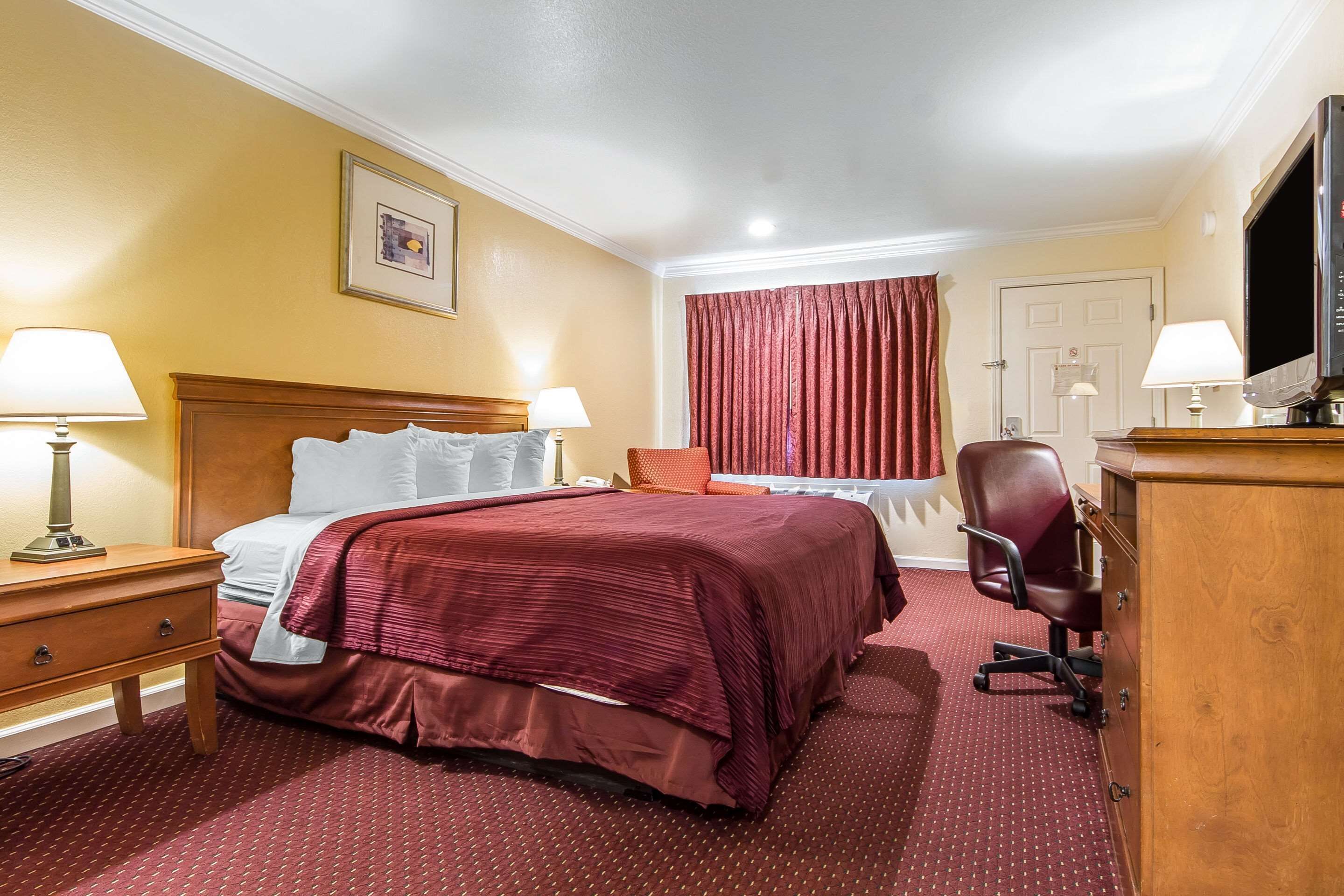 A hotel room with a large bed, two bedside tables with lamps, a wooden dresser, a TV, and a red curtain.