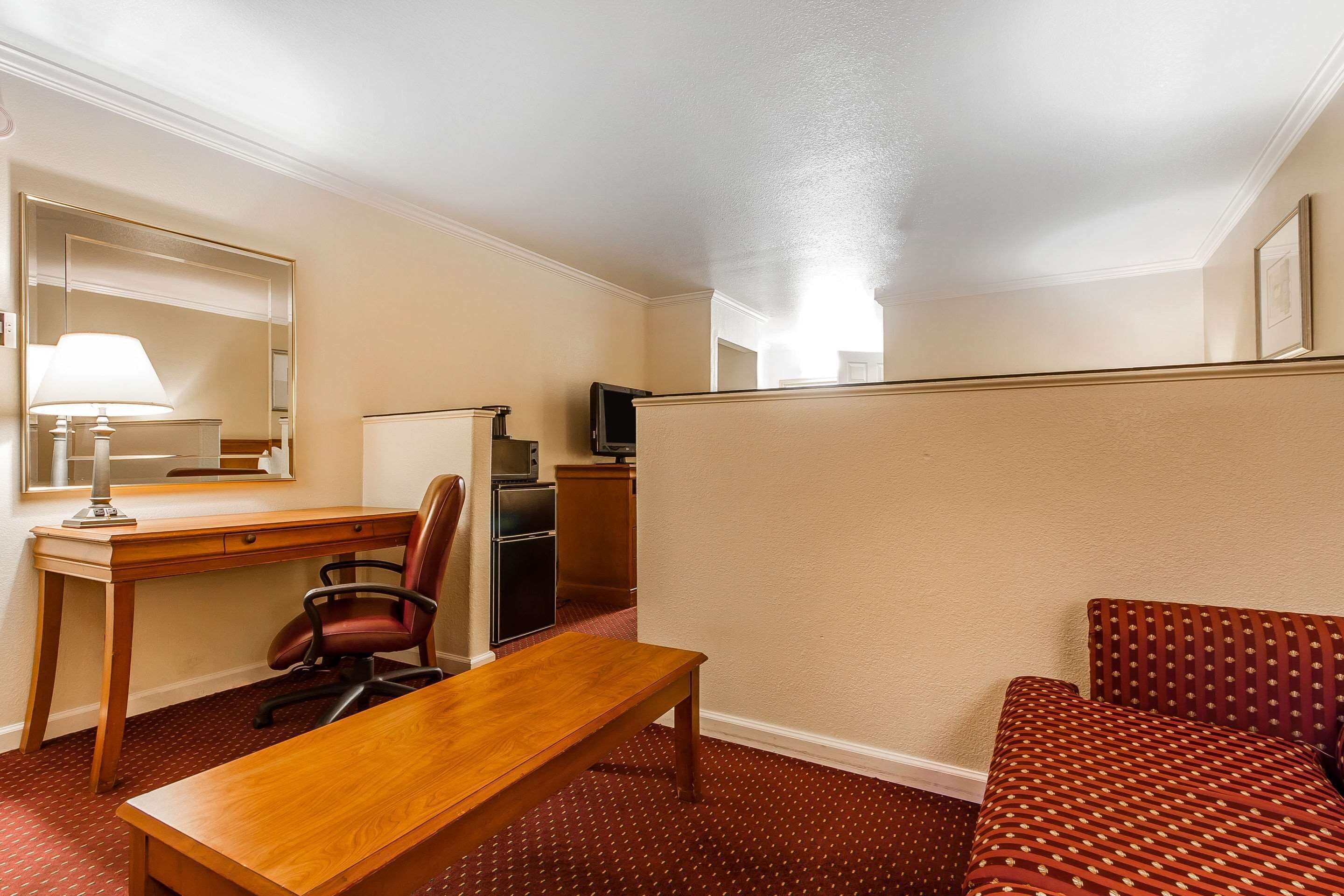 A hotel room with a wooden desk, a chair, a bench, a television, and a patterned sofa.