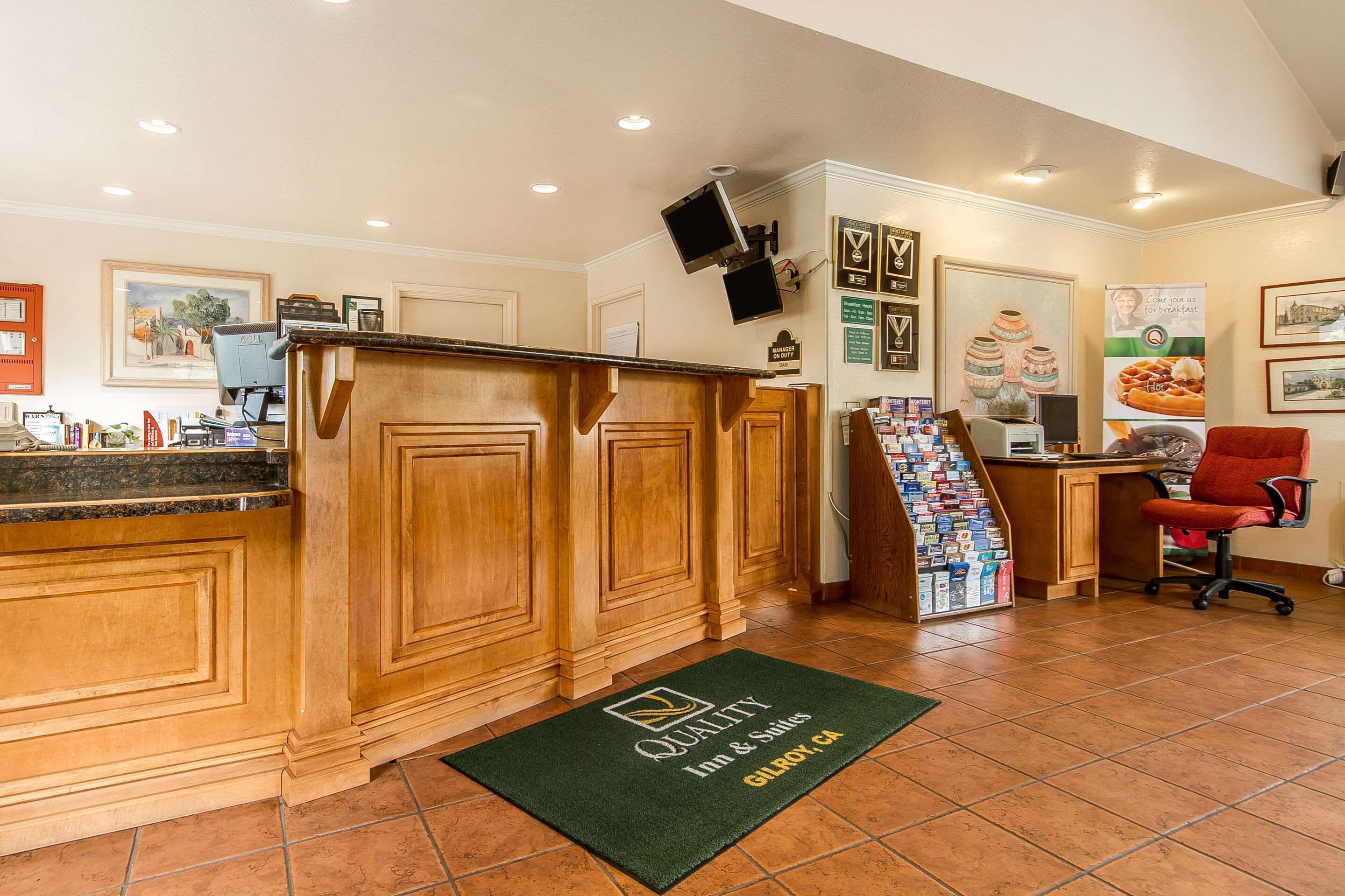 A reception area with a wooden counter, a computer, and a desk with a chair. There is a green doormat with the hotel name and location.