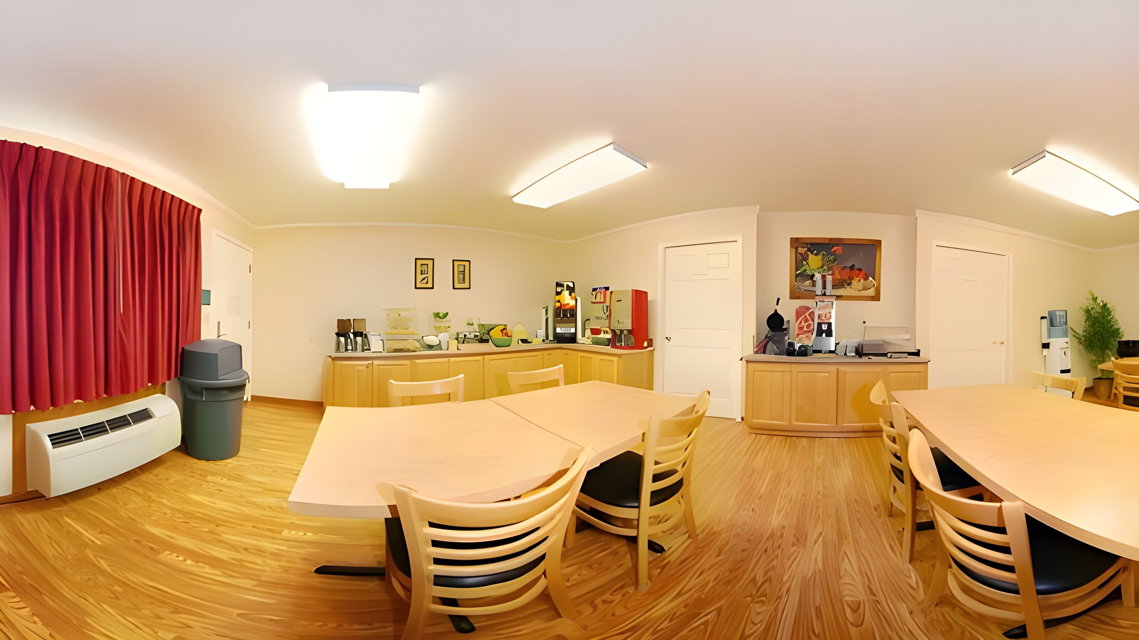 A compact, well-lit room with a dining table, chairs, a kitchen area, and a trash bin.