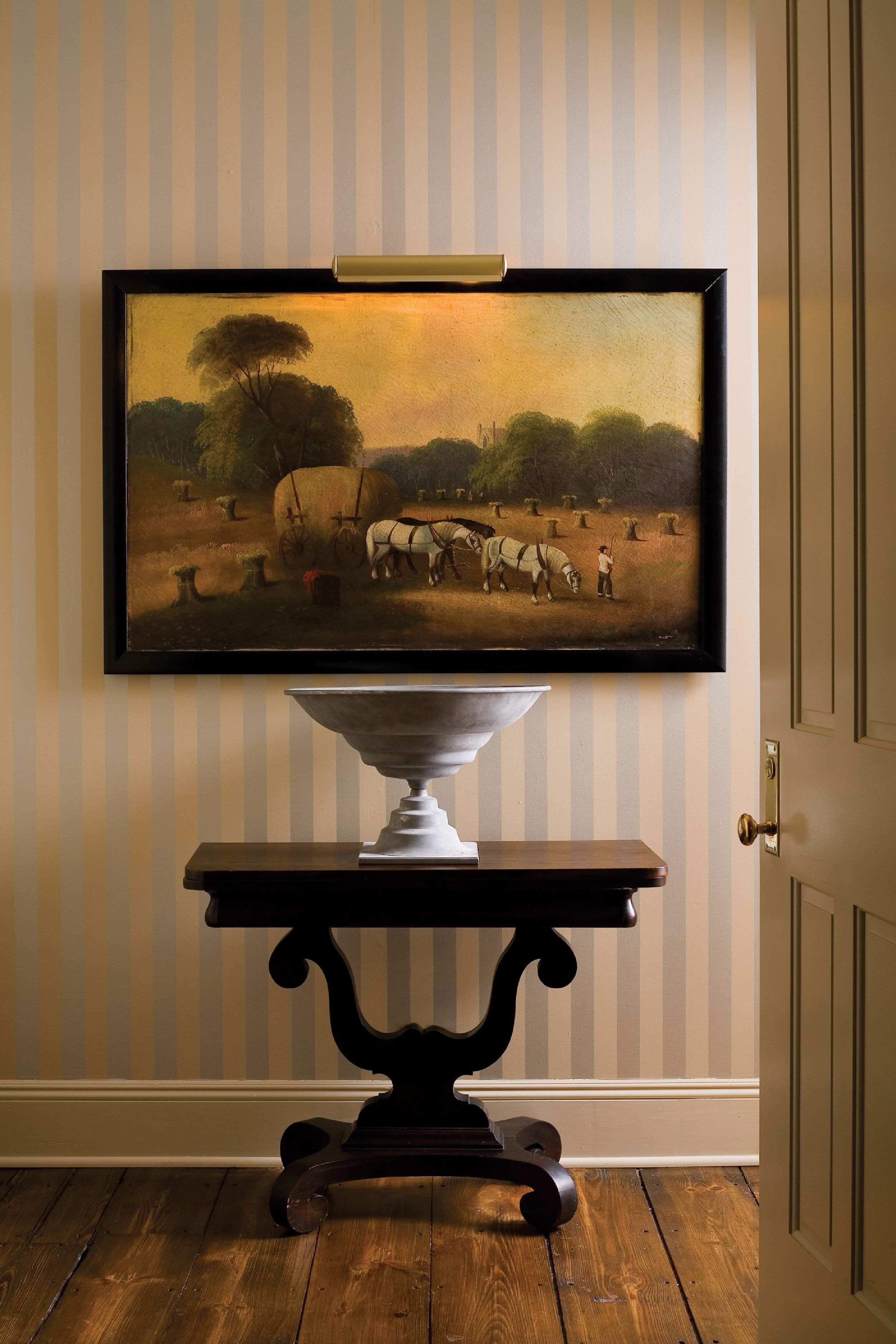 Un cuadro enmarco de una escena campestre con hileras de pacas de heno y un carruaje tirado por caballos, colgado en una pared sobre una mesa pedestal blanca con base de madera oscura.
