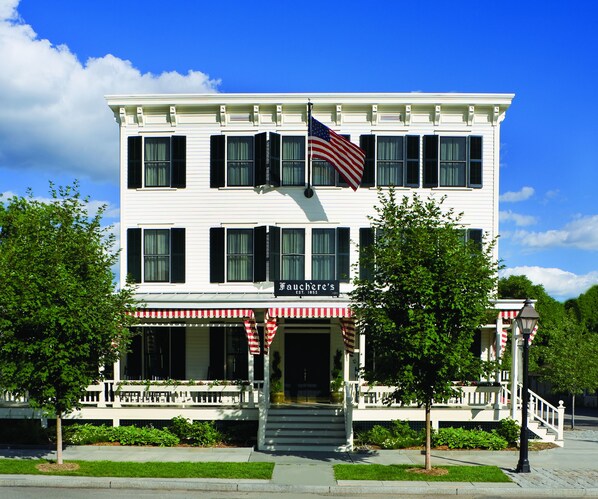 A two-story building with a white facade, black shutters, and a red and white striped awning.