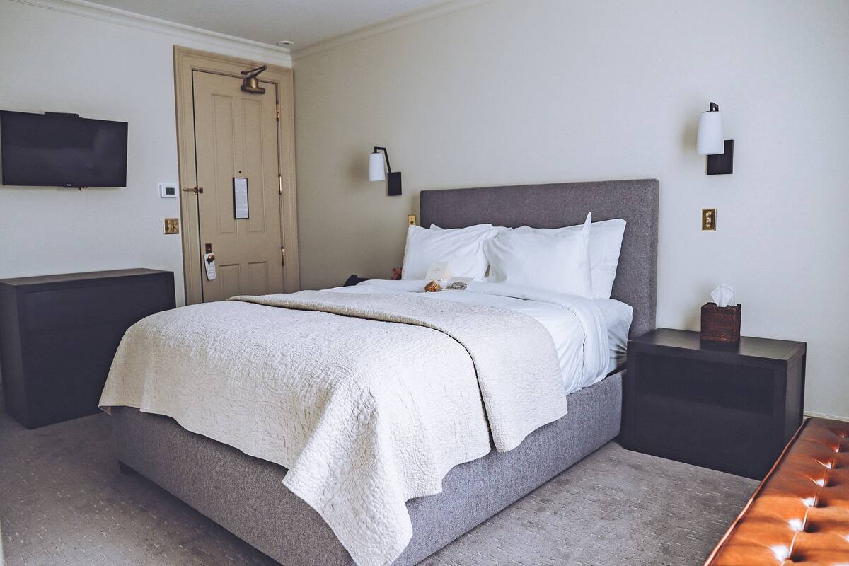 A neatly made bed with a white comforter and pillows, a bedside table with a lamp, and a flat-screen TV mounted on the wall.