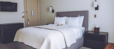 Ein ordentlich bezogenes Bett mit weißer Decke und Kissen, ein Nachttisch mit Lampe sowie ein an der Wand befestigter Flachbildfernseher.