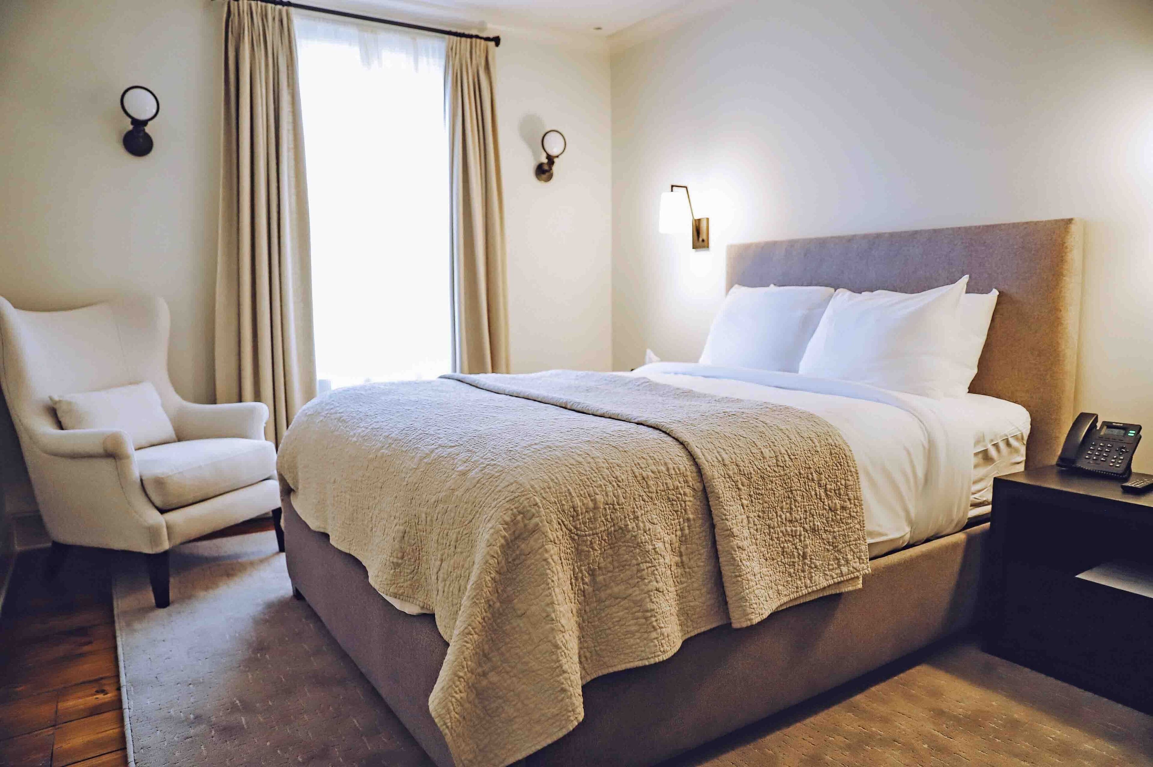 Una habitación de hotel con una cama grande, una silla, una mesita de noche con teléfono y una ventana con cortinas.