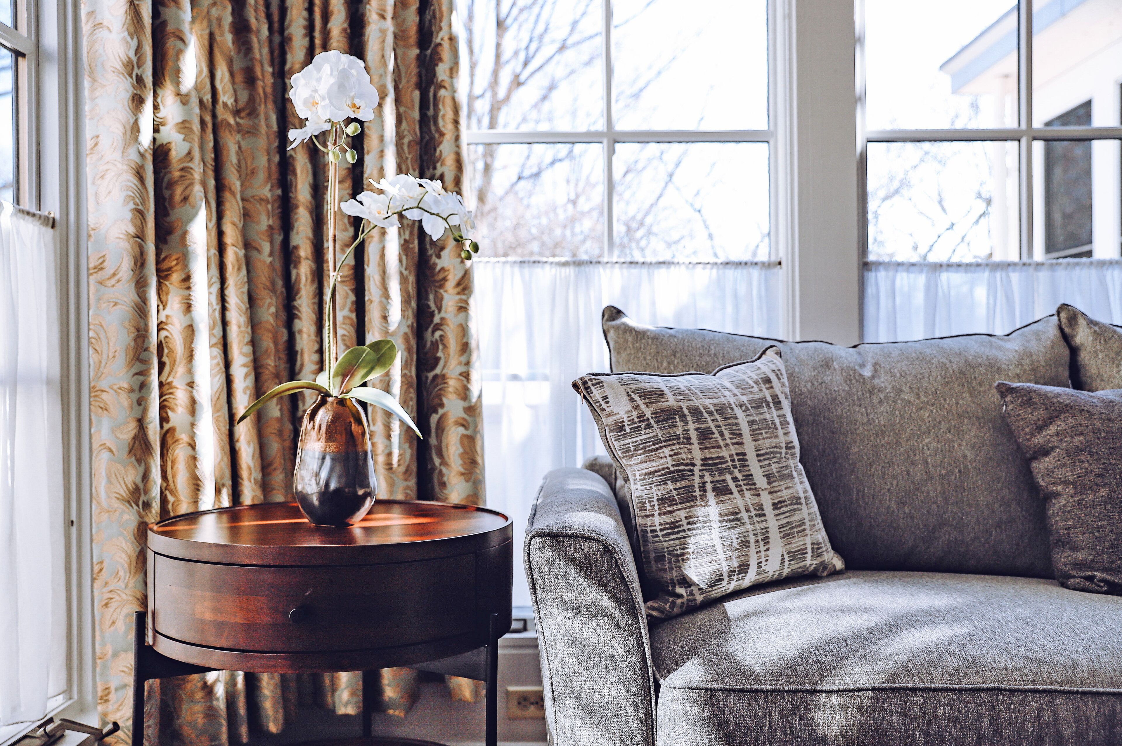 A living room with a grey sofa, a patterned throw pillow, and a vase with white flowers on a wooden side table.